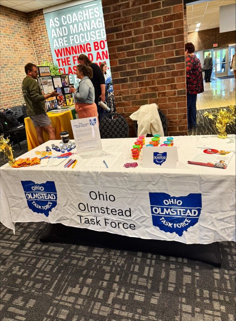 Picture of conference table with Ohio Olmstead Task Force banner and swag.  People talking and brick walls in the background.