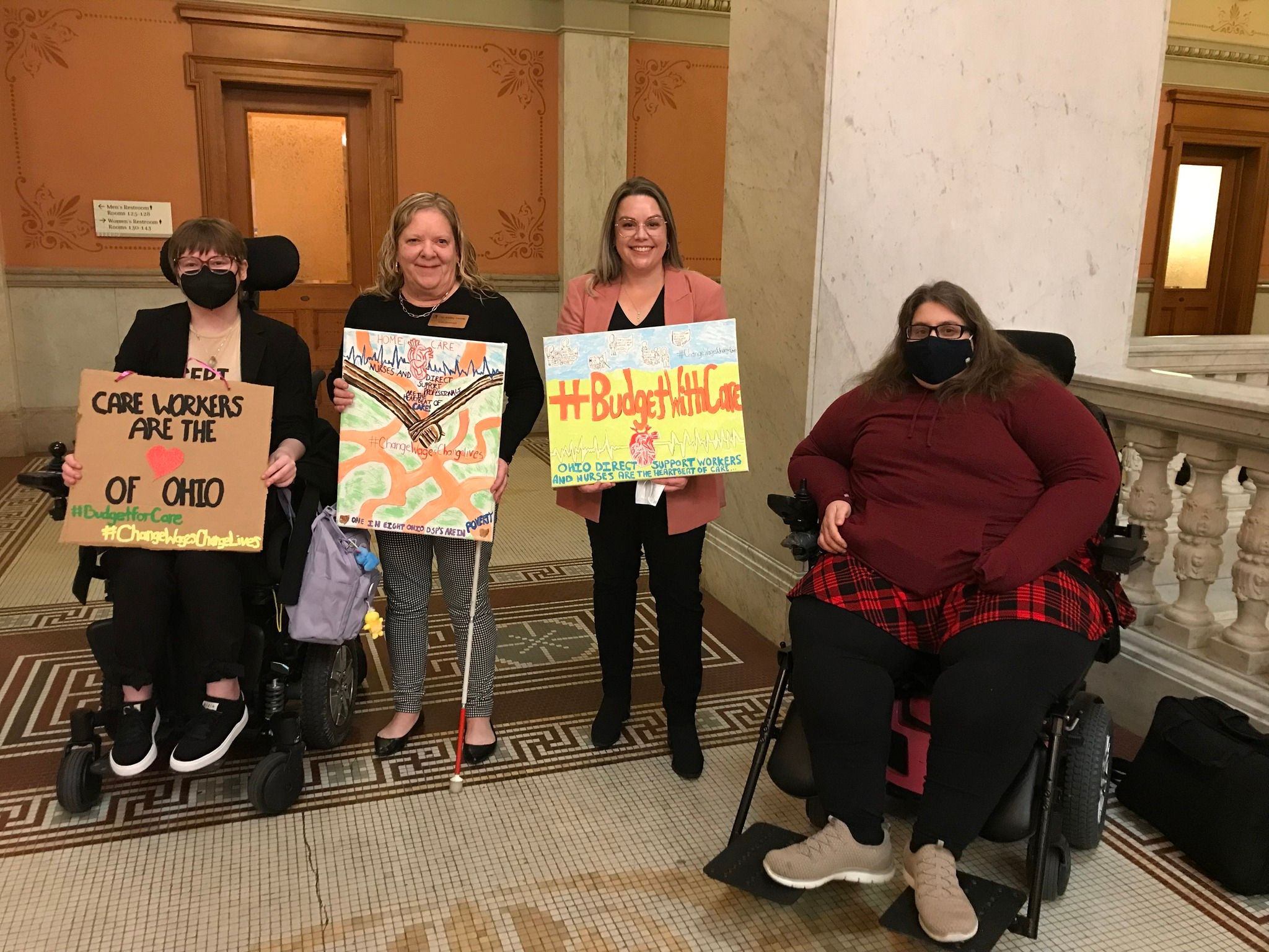 Four OOTF advocates sit and stand with homemade signs supporting direct support workers and carers at the Ohio Statehouse