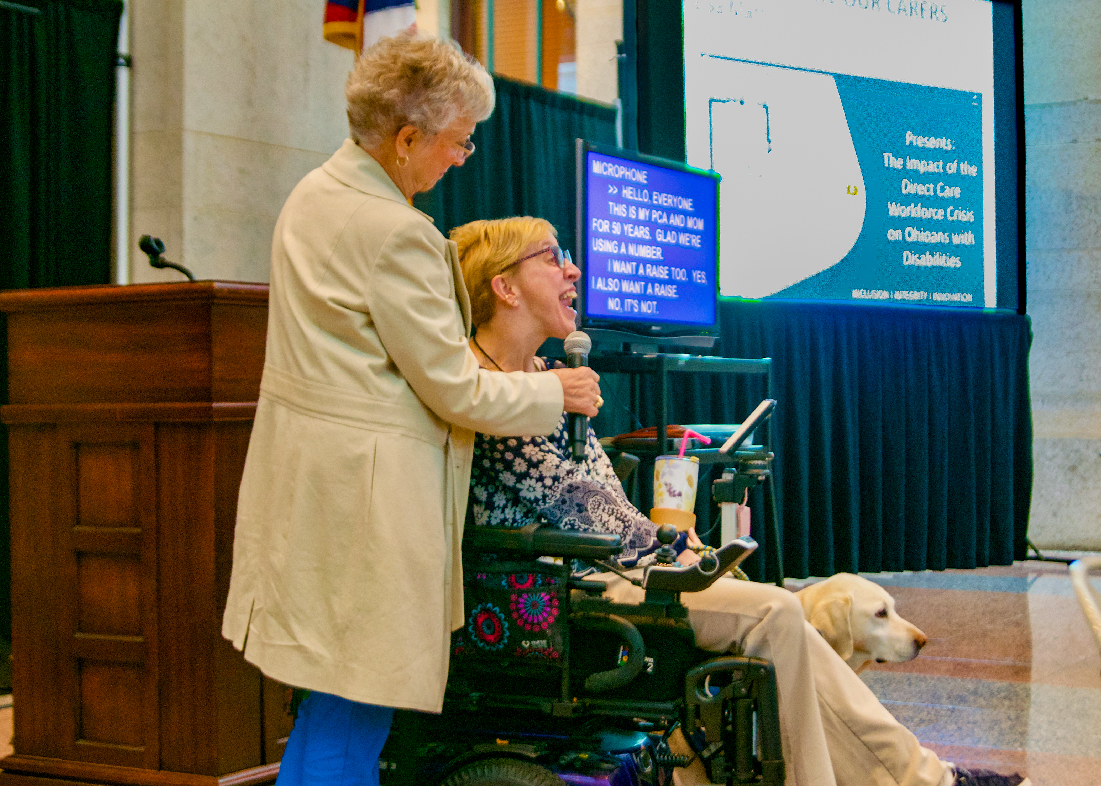 OOTF advocate speaks into microphone about her support for direct care workers. Another woman is holding the microphone to her mouth, and her service dog is behind her wheelchair.